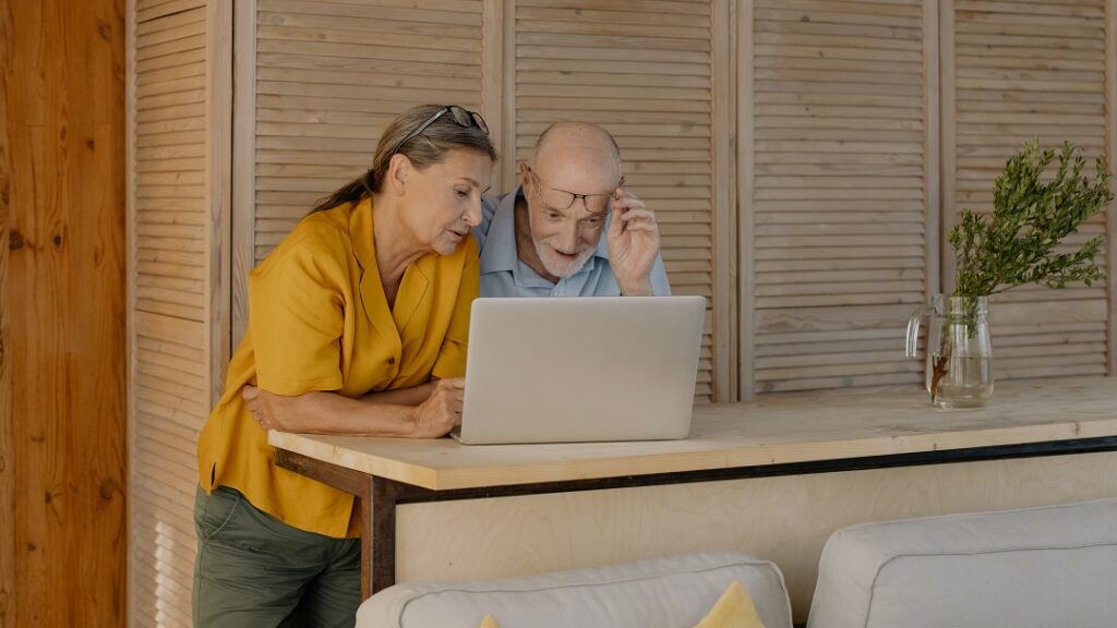 Een man een vrouw van rond de zestig staan kijken samen op een laptopscherm. De man doet zijn bril af om het te kunnen zien.