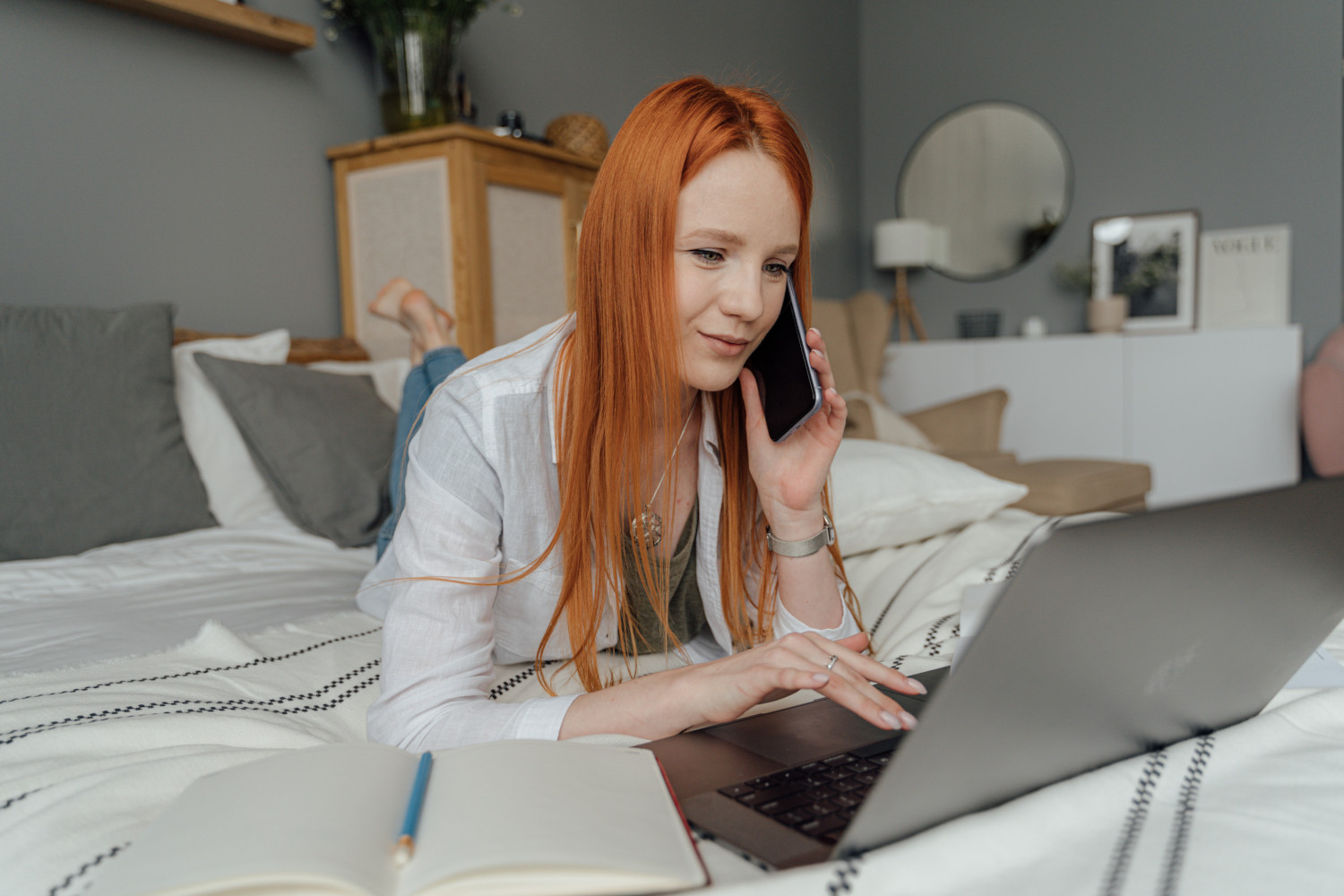 Een vrouw ligt op bed met haar laptop en belt met haar telefoon in haar linkerhand