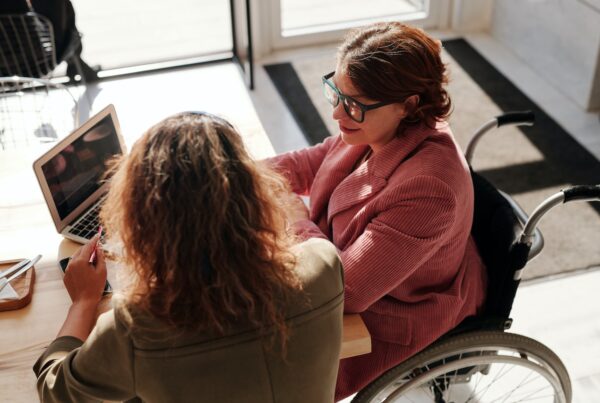 vrouw in rolstoel zit aan tafel met een laptop en praat met een andere vrouw