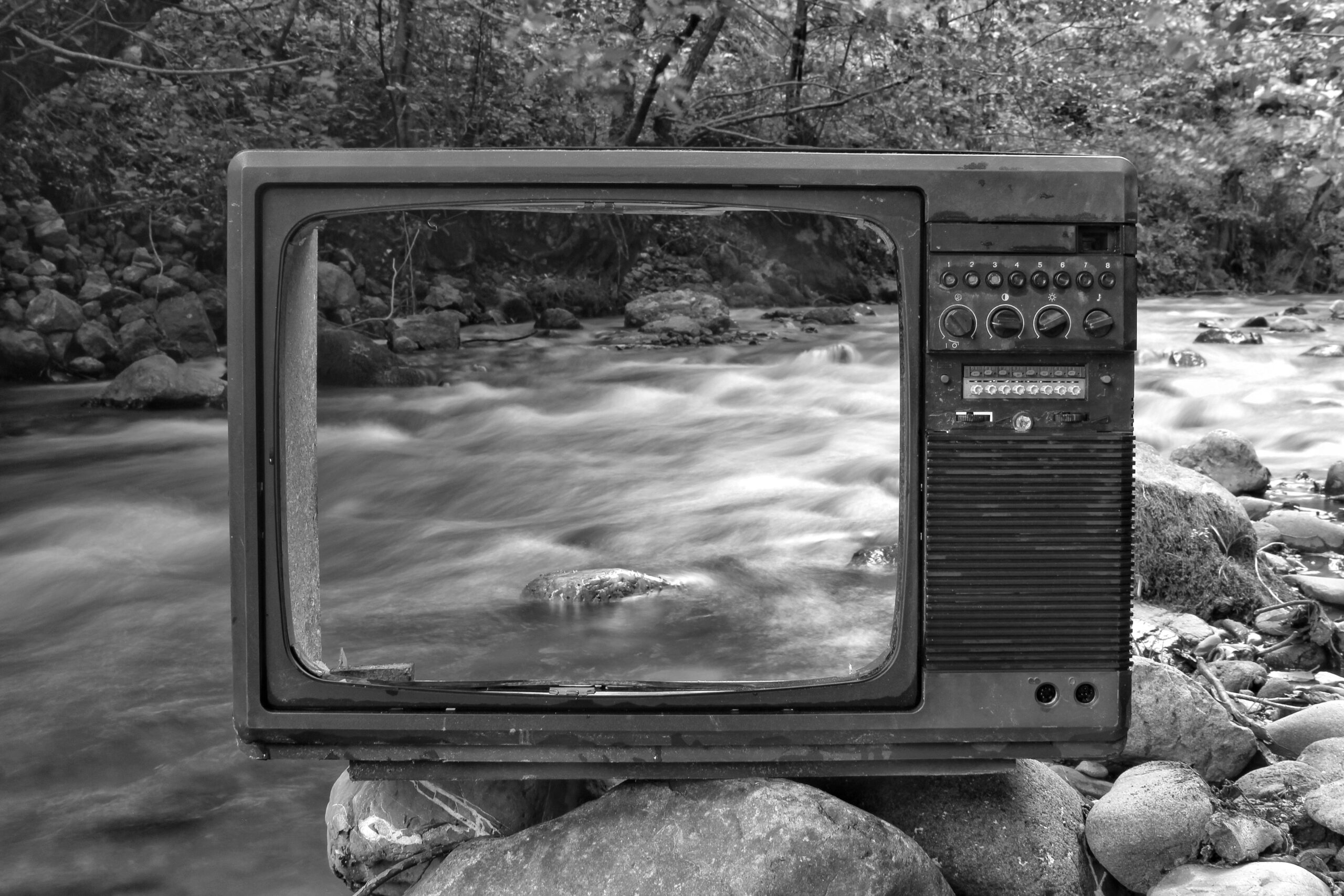 Deze artistieke zwartwit-foto laat het geraamte zien van een oud tv-toestel, op grote keien, aan de oever van een stromend beekje.