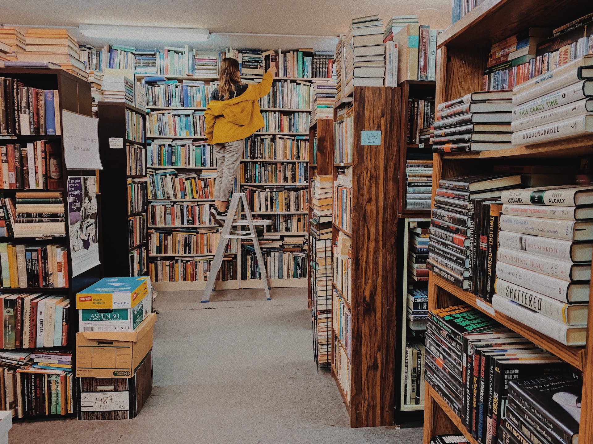 in een boekenwinkel staat een vrouw met een gele jas op een ladder om een boek te pakken