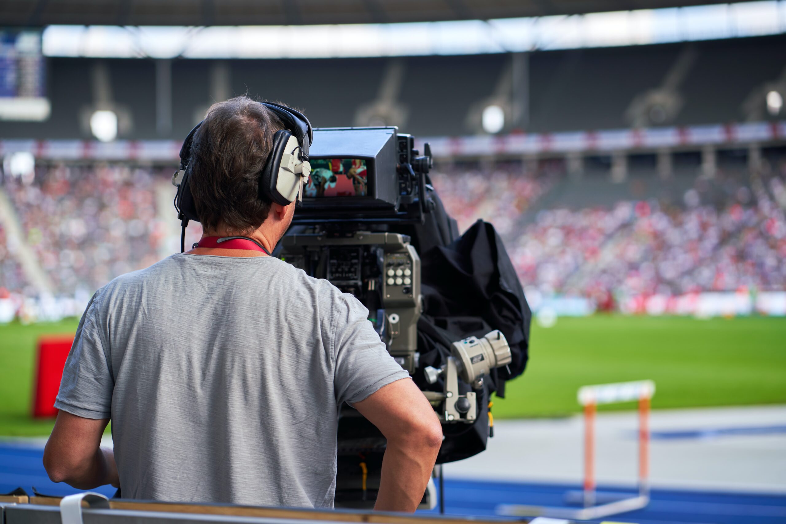 We zien het bovenlijf van een cameraman in een grijs t-shirt van achteren gefotografeerd, terwijl hij een sportevenement in een stadion filmt.